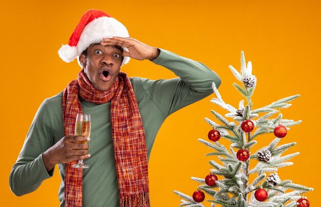 African american man in santa hat and scarf around neck holding glass of champagne looking far away surprised with hand over head standing next to a christmas tree over orange background