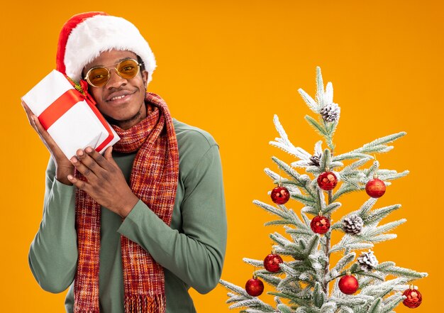 Uomo afroamericano in santa cappello e sciarpa intorno al collo tenendo un presente guardando la fotocamera con il sorriso sul viso in piedi accanto a un albero di natale su sfondo arancione