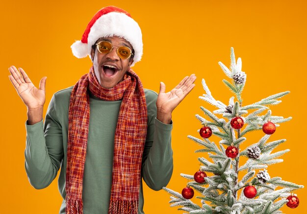 African american man in santa hat and scarf around neck looking at camera happy and cheerful standing next to a christmas tree over orange background