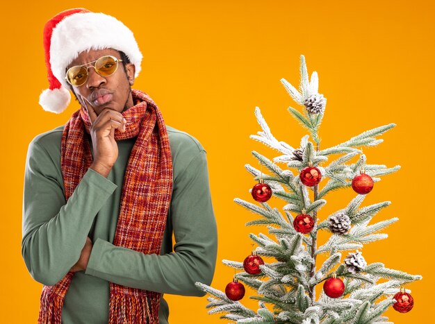 African american man in santa hat and scarf around neck looking at camera with pensive expression thinking standing next to a christmas tree over orange background