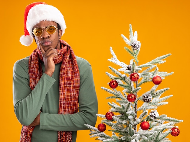 African american man in santa hat and scarf around neck looking at camera with pensive expression thinking standing next to a christmas tree over orange background