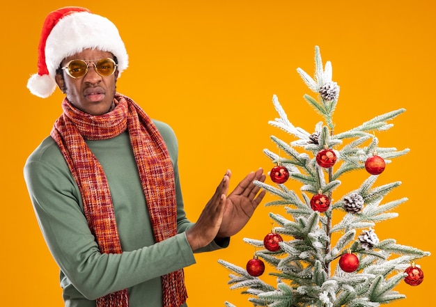 Free photo african american man in santa hat and scarf around neck looking displeased making defense gesture standing next to a christmas tree over orange background