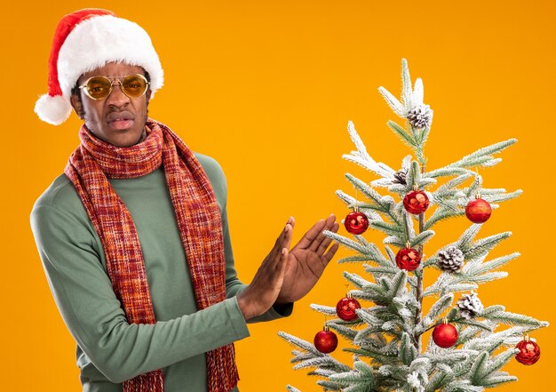산타 모자와 스카프 목에 아프리카 계 미국인 남자 오렌지 배경 위에 크리스마스 트리 옆에 서있는 방어 제스처를 만드는 불쾌감을 찾고