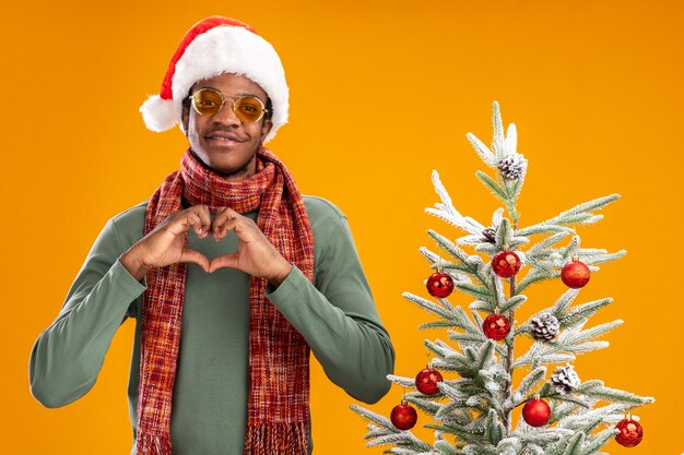 オレンジ色の背景の上のクリスマスツリーの横に立って笑っている指でハートジェスチャーを作る首の周りのサンタの帽子とスカーフのアフリカ系アメリカ人の男