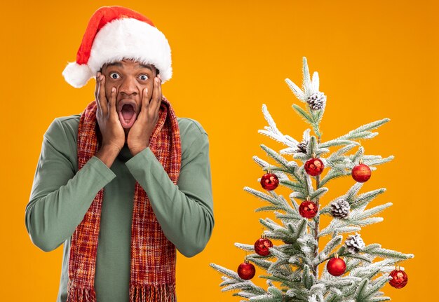 African american man in santa hat and scarf around neck  shocked standing next to a christmas tree over orange wall