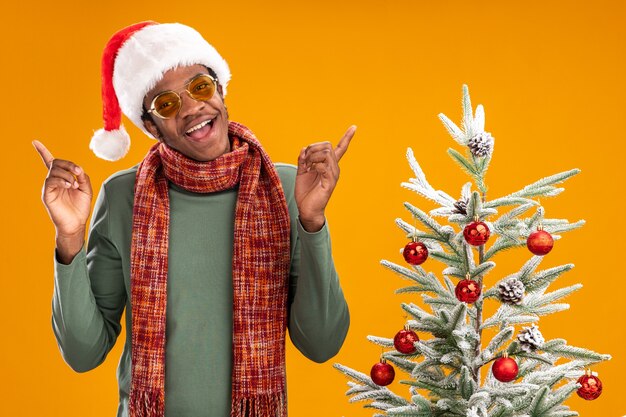 African american man in santa hat and scarf around neck  smiling cheerfully standing next to a christmas tree over orange wall