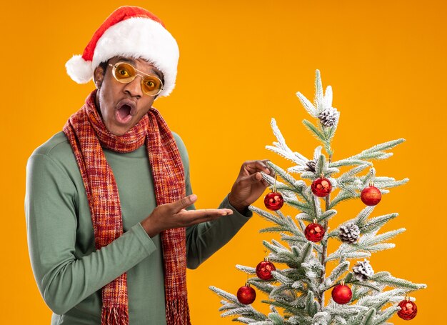 African american man in santa hat and scarf around neck standing next to a christmas tree presenting with arms looking surprised over orange background