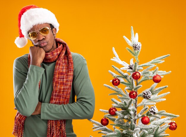 African american man in santa hat and scarf around neck  with pensive expression on face thinking standing next to a christmas tree over orange wall