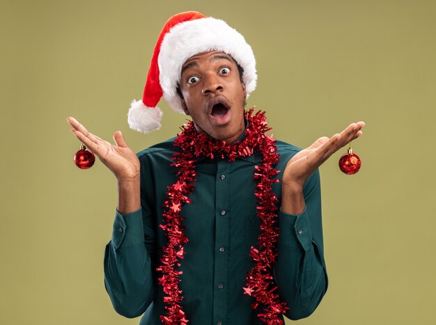 African american man in santa hat with garland holding christmas balls looking at camera amazed and surprised standing over green background