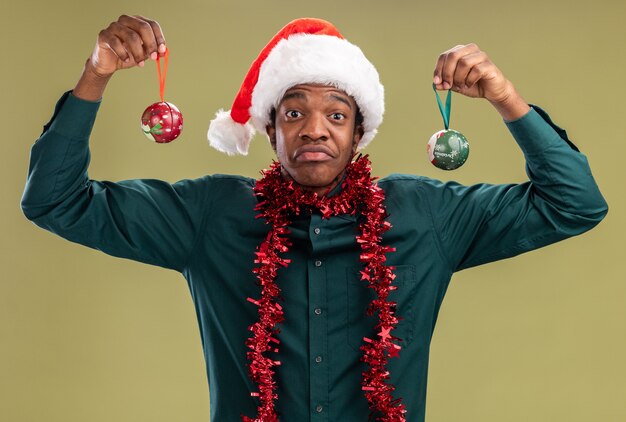 크리스마스 공을 들고 갈 랜드와 산타 모자에 아프리카 계 미국인 남자 놀라게 하 고 녹색 벽 위에 서 혼란