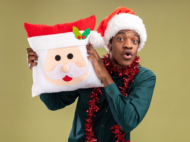 緑の背景の上に立って驚いたカメラを見てクリスマス枕を保持している花輪とサンタ帽子のアフリカ系アメリカ人の男