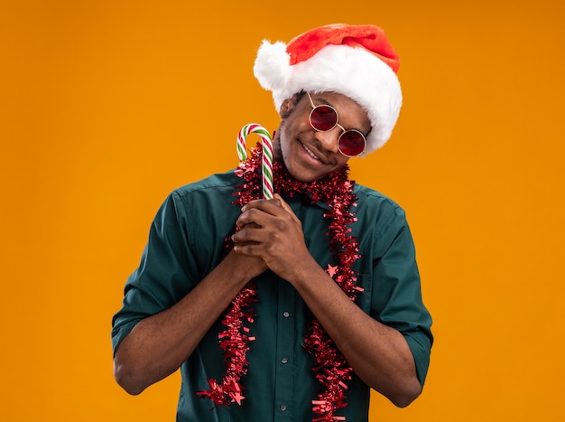 Free photo african american man in santa hat with garland wearing glasses holding candy cane looking at camera with smile on face standing over orange background