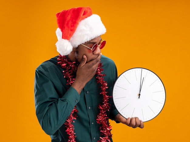 Uomo afroamericano in cappello della santa con la ghirlanda che indossa gli occhiali da sole che tengono l'orologio guardandolo stupito e sorpreso in piedi su sfondo arancione