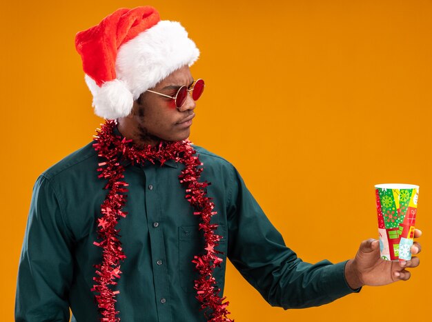 Uomo afroamericano in cappello della santa con la ghirlanda che indossa gli occhiali da sole che tengono il bicchiere di carta colorato guardandolo con la faccia seria che sta sopra la parete arancione