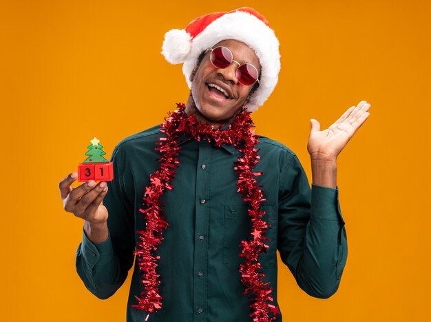 African american man in santa hat with garland wearing sunglasses holding toy cubes with new year date happy and cheerful with raised arm standing over orange wall