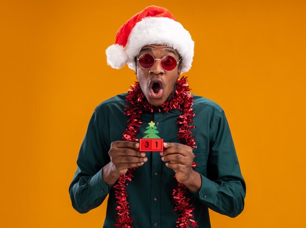 African american man in santa hat with garland wearing sunglasses holding toy cubes with new year date looking at camera amazed and shocked standing over orange background