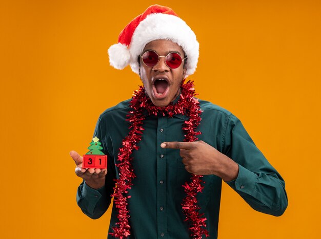 African american man in santa hat with garland wearing sunglasses holding toy cubes with new year date pointing with index finger at it surprised and amazed standing over orange background