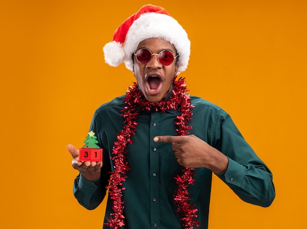 African american man in santa hat with garland wearing sunglasses holding toy cubes with new year date pointing with index finger at it surprised and amazed standing over orange background