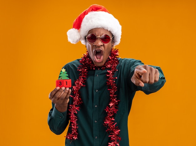 African american man in santa hat with garland wearing sunglasses holding toy cubes with new year date shouting with aggressive expression pointing with index finger standing