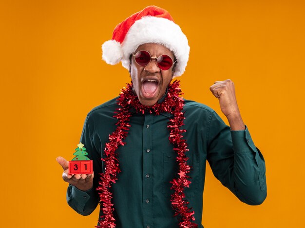 오렌지 벽 위에 서있는 공격적인 표현으로 외치는 새해 날짜와 장난감 큐브를 들고 선글라스를 착용하는 갈 랜드와 산타 모자에 아프리카 계 미국인 남자