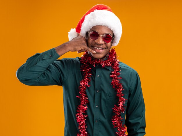 African american man in santa hat with garland wearing sunglasses  smiling cheerfully making call me gesture standing over orange wall