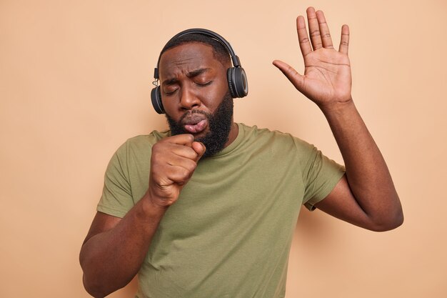 Афроамериканец поет песню, держит руку у рта, как будто микрофон слушает музыку из плеера, использует беспроводные наушники, носит повседневную футболку, развлекается в свободное время, изолированно от бежевой стены