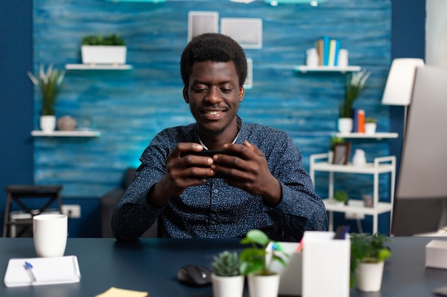 African american man smiling while using a smartphone at desk to check social media remote working g...