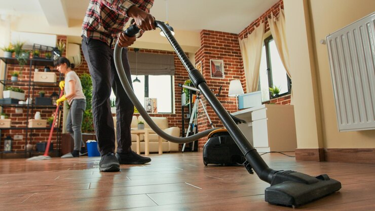 Free Photo african american man vacuuming floors in living room, cleaning apartment with girlfriend. young adult using vacuum cleaner and woman wiping shelves with all purpose cleaner. tripod shot.