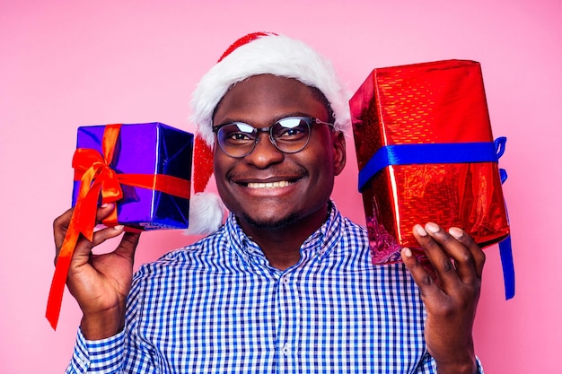 세련된 격자 무늬 셔츠를 입은 아프리카 계 미국인 남자는 분홍색 배경 studio.dark-skinned 산타 클로스 메리 크리스마스를 축하하는 선물 상자와 산타 모자에 큰 미소