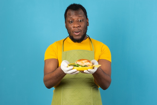 African American man with beard, wearing apron with a yellow plate of sandwich in hands , stands over blue wall. 