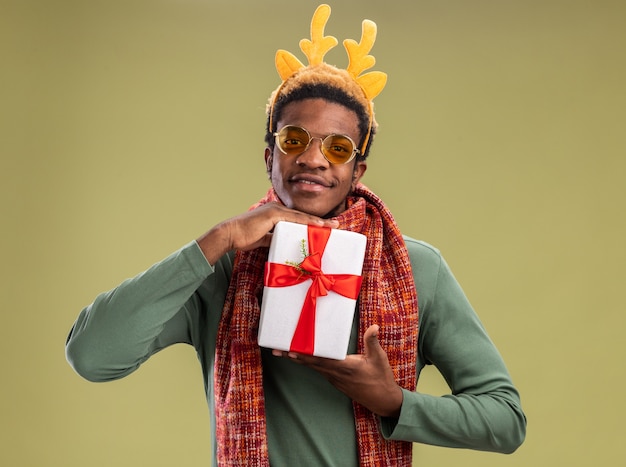 Uomo afroamericano con bordo divertente con corna di cervo e sciarpa intorno al collo tenendo presente il regalo di natale guardando la fotocamera con il sorriso sul viso in piedi su sfondo verde