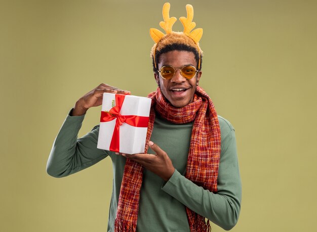 African american man with funny rim with deer horns and scarf around neck holding christmas present looking at camera with smile on face standing over green background