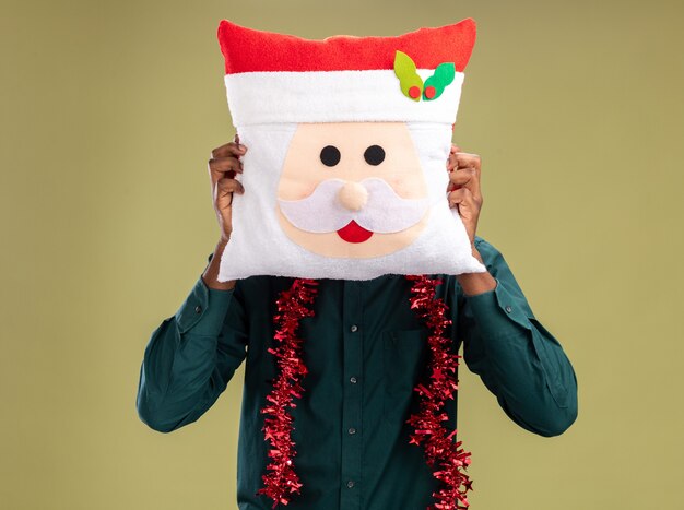 African american man with garland holding christmas pillow hiding his face behind pillow standing over green background