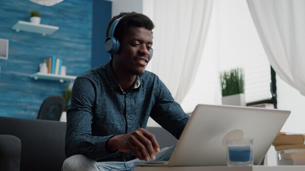 Афро-американский мужчина с наушниками смотрит фильм на потоковых сервисах в яркой гостиной, пользователь компьютера сосредоточился на офисной работе или домашних развлечениях, улыбается