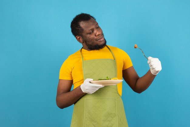 African American man with meal in one hand and fork in the other , stands over blue wall. 