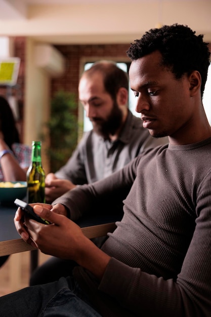 無料写真 インターネットを閲覧しながらテーブルに座っている現代のタッチ スクリーン スマート フォンを持つアフリカ系アメリカ人男性。ブラウザとソーシャル メディア フィードをナビゲートしながら自宅の居間に座っている人。
