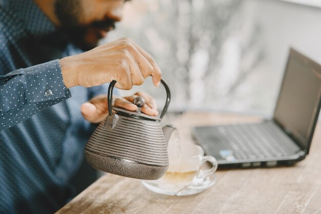 African-american man working behind a laptop and writing in a notebook. Man with beard sitting in a cafe and pouring a tea.