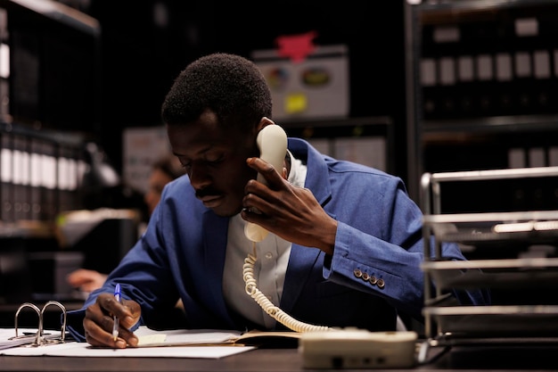 Free photo african american manager talking at landline phone, discussing management research with remote bookkeeper in storage room. businessman working late at night at accountancy report