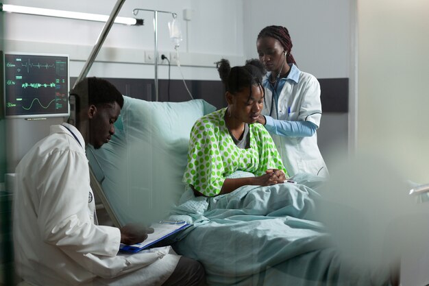 African american medical team working on healing patient in hospital ward bed. Man and woman with doctors occupation examining young adult for treatment using monitor and stethoscope