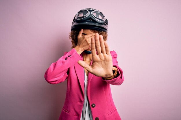 African american motorcyclist woman with curly hair moto helmet over pink background covering eyes