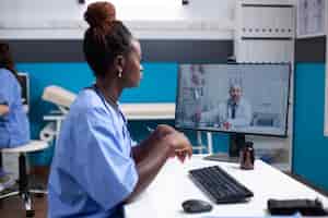 Free photo african american nurse at clinic desk listening to general practitioner collegue doctor during video conferencing. medical team in telemedicine call at busy modern hospital office