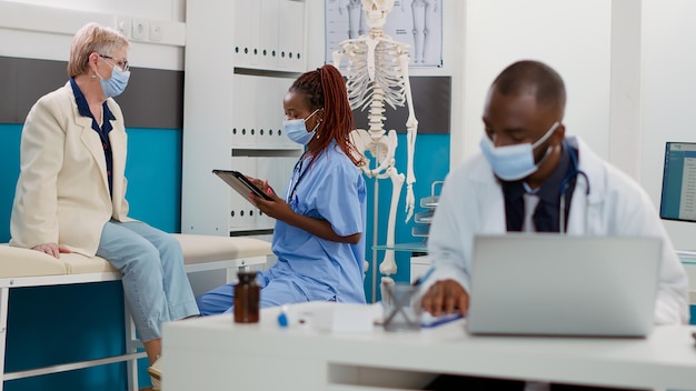 Афро-американская медсестра консультирует пожилого пациента с помощью планшета, чтобы делать заметки, проводит осмотр во время пандемии ковида 19. Специалист и пожилая женщина на приеме у врача.