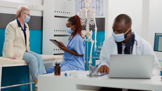 African american nurse consulting senior patient using tablet to take notes, doing examination during covid 19 pandemic. Specialist and elderly woman having checkup visit appointment.