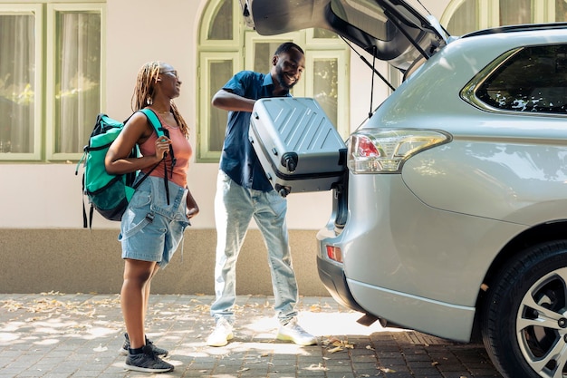 アフリカ系アメリカ人のパートナーが車で旅行し、荷物や荷物をトランクに入れて夏休みに出発します。トロリーやトラベルバッグを持って休暇旅行に行く関係の人々。