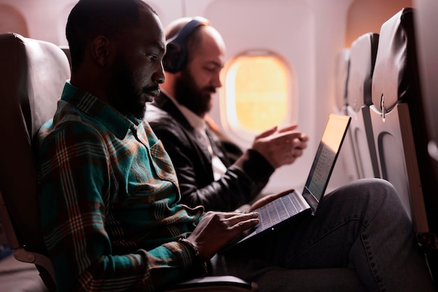 아프리카계 미국인 승객은 일몰 비행 중에 노트북 작업을 하고 휴가 목적지에 도착하기를 기다리고 있습니다. 온라인으로 컴퓨터를 사용하여 관광객 그룹과 이코노미 클래스로 비행합니다.