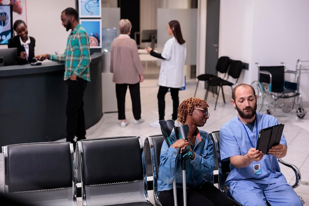 African american patient and nurse looking at tablet to find dsease diagnosis on digital device, suffering from physical impairment in waiting area. Woman and specialist in hospital lobby.