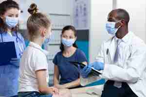 Free photo african american pediatrician doctor with protection face mask against coronavirus explaining medical treatment to family during clinical appointment in hospital office. health care service