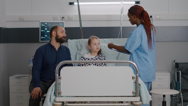 Foto gratuita infermiera pediatra afroamericana che controlla la temperatura utilizzando un termometro a infrarossi che monitora i sintomi della malattia durante l'esame sanitario nel reparto ospedaliero. assistente alla scrittura di perizie mediche