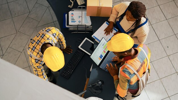 African american people looking at supplies list on pc, working with computer to check inventory and logistics. Employees organizing merchandise on shelves in warehouse space. Handheld shot.