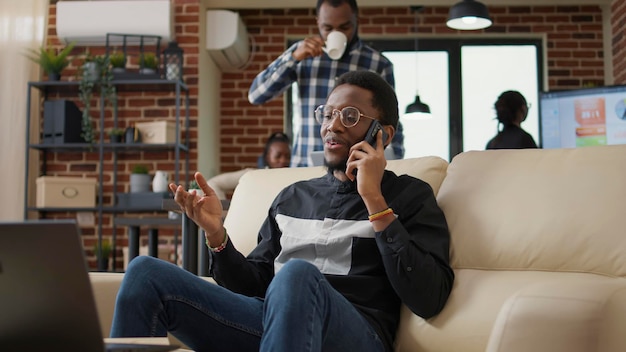 African american person chatting on phone call in business office, using smartphone to enjoy remote telecommunication with colleagues. Young man working on laptop and having conversation.